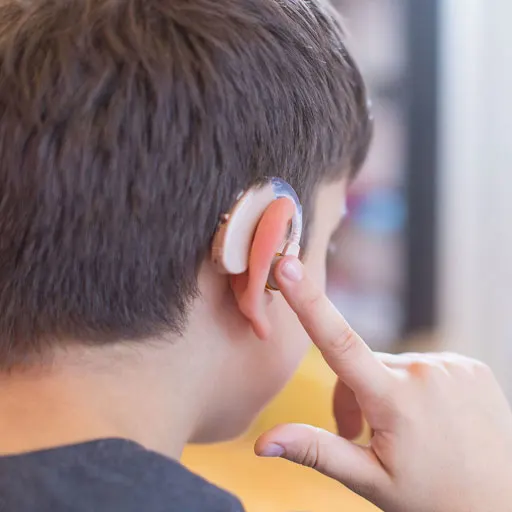 oung boy wearing a skin-colored hearing aid; he's tapping it to adjust or control sound settings.